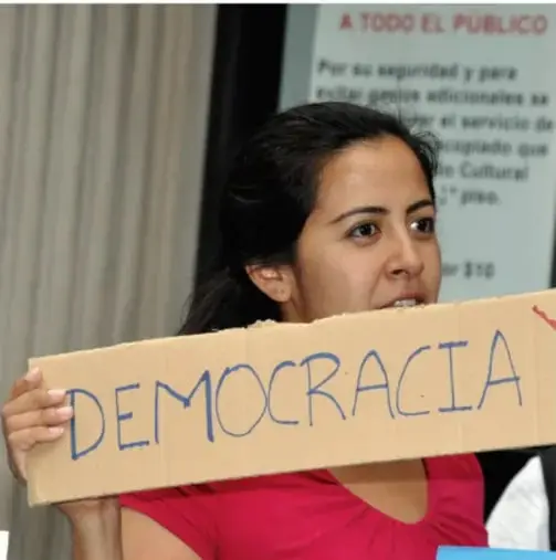 Fotografía de una mujer joven sosteniendo un cartel de cartón con la palabra DEMOCRACIA en letras azules, en un acto público.
