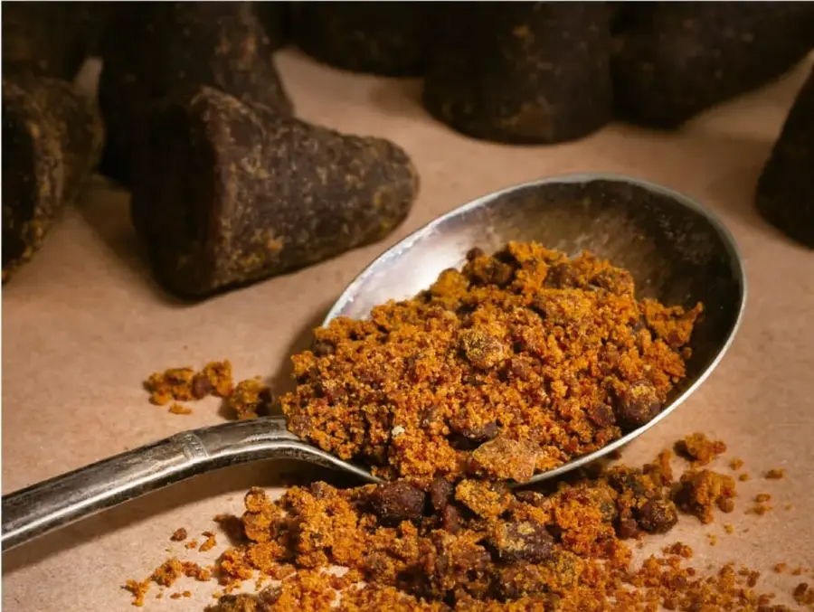 Fotografía: cuchara de metal sobre superficie clara con un montón de granulado o polvo de color café anaranjado; alrededor hay piezas macizas de color café muy oscuro (posiblemente chocolate o fruta deshidratada), ilustrando un ejemplo de producción con innovación y menor impacto ambiental.