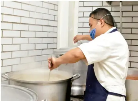 Fotografía de un cocinero con cofia y mandil, removiendo un gran recipiente metálico en una cocina de paredes de ladrillo blanco.