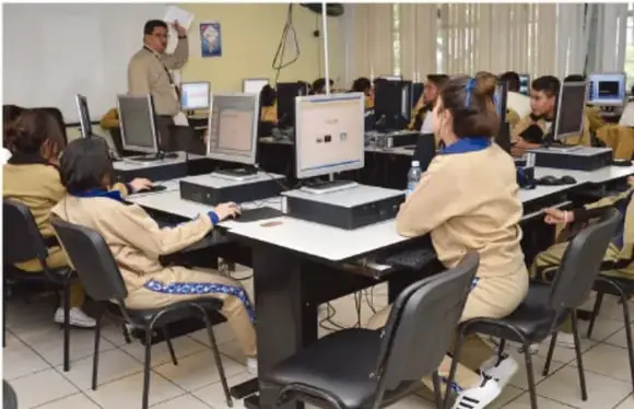Fotografía de un laboratorio de cómputo escolar: un docente de pie frente a estudiantes sentados que trabajan con computadoras de escritorio.