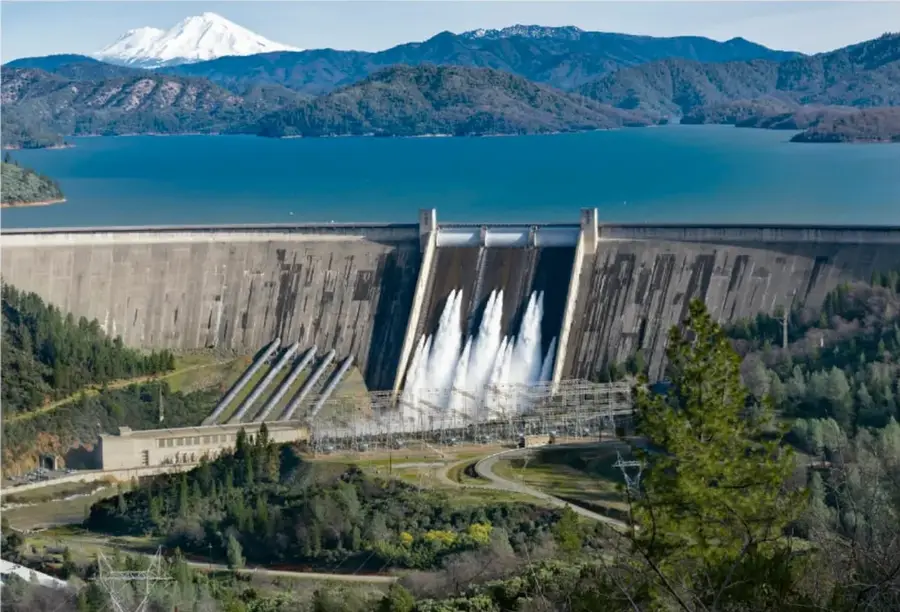 Fotografía de una presa de concreto con compuertas abiertas descargando agua, un embalse azul, montañas boscosas alrededor y un volcán nevado al fondo.