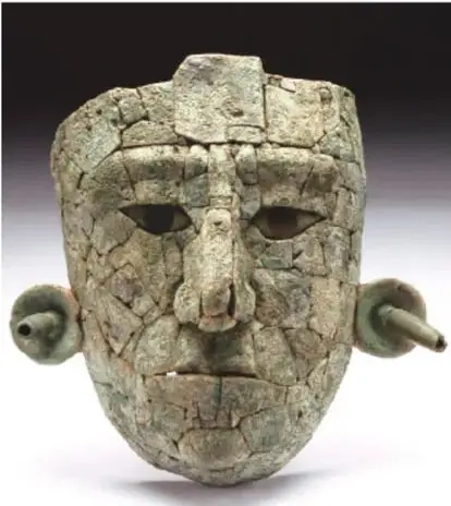 Fotografía de una máscara antigua de rostro humano elaborada en piezas de piedra o jade ensambladas, con ojos almendrados, nariz prominente, orejeras circulares y grietas visibles; corresponde a una máscara mesoamericana de estilo Preclásico/Clásico.