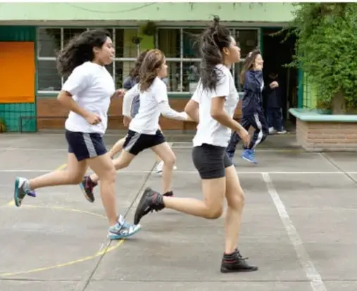 Fotografía de tres estudiantes adolescentes con uniforme blanco y pantalón oscuro corriendo en el patio de una escuela, realizando actividad física al aire libre.
