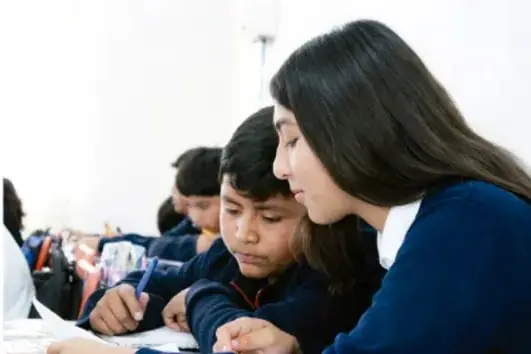 Fotografía de una docente apoyando a un estudiante que escribe en su cuaderno, con otros alumnos trabajando alrededor en el aula.