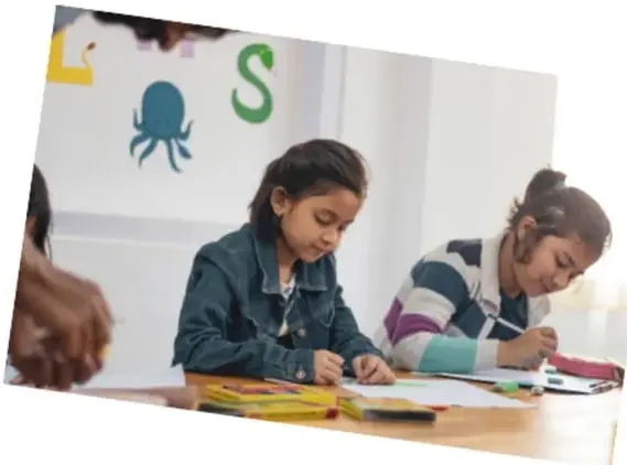 Una niña escribe en una hoja mientras una maestra la observa y orienta; al fondo otros estudiantes trabajan.