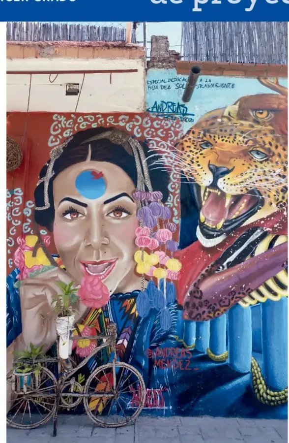 Mural fotografiado en una calle: retrato de una mujer con rasgos inspirados en Frida Kahlo, con cintas y flores en el cabello, y a su lado un jaguar o leopardo rugiente de colores naranjas y azules; al frente, una bicicleta artesanal con macetas de plantas.
