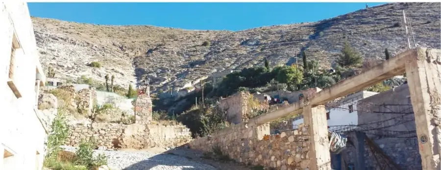 Panorámica de un cañón con muros de piedra, construcciones antiguas, árboles y cerros pelados bajo cielo azul.
