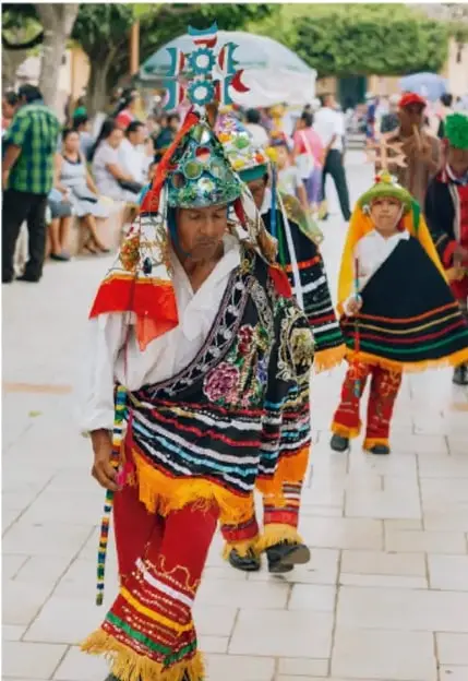 Fotografía de personas (adultos y niños) con trajes típicos indígenas muy coloridos, portando objetos ceremoniales, participando en una celebración o desfile en la vía pública, con público observando de fondo.