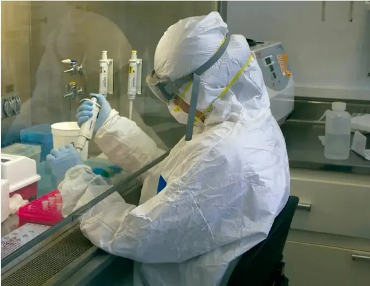 Fotografía de una científica o técnico de laboratorio cubierto con traje blanco de protección total, careta y guantes, trabajando dentro de una campana de flujo laminar; manipula una pipeta y tubos de ensayo con muestra roja, en un entorno de laboratorio con material de vidrio y frascos.