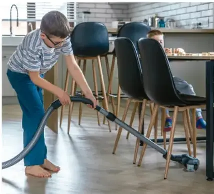 Niño aspirando el piso de una cocina con una aspiradora, mientras una mujer está sentada al fondo.