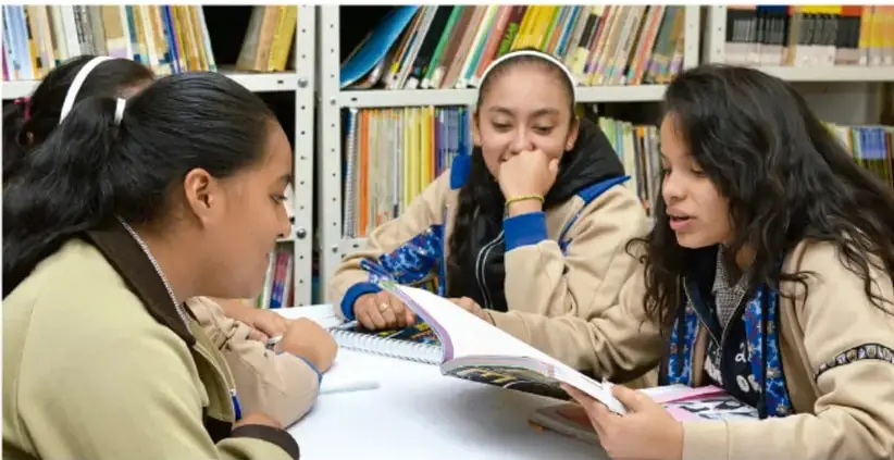 Fotografía de tres estudiantes en una biblioteca, sentadas alrededor de una mesa, revisando juntas un libro y un cuaderno; al fondo hay estantes con libros.