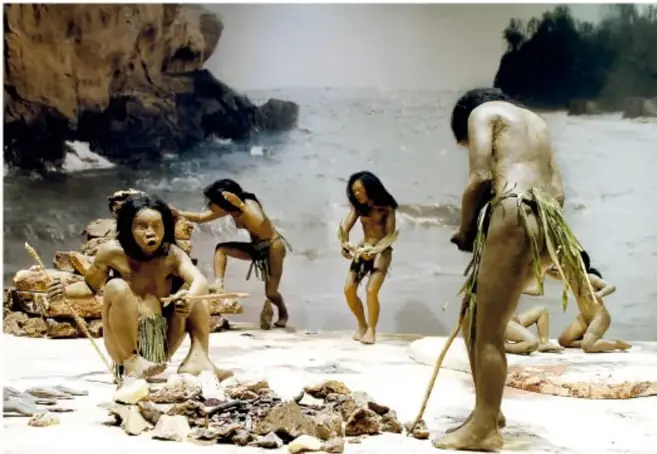 Maqueta de un grupo de personas de la prehistoria junto a una fogata en la playa, algunas de pie con varas y piedras en el suelo, la caza o pesca del día.