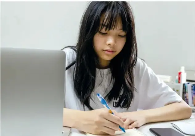 Fotografía de una estudiante sentada en un escritorio, con una laptop abierta, escribiendo en un cuaderno con un bolígrafo; representa el trabajo individual de autoevaluación.