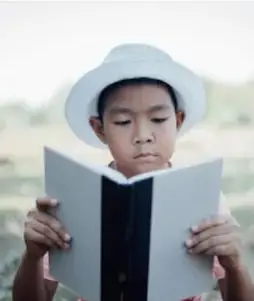 Fotografía de un niño con sombrero leyendo un libro al aire libre.