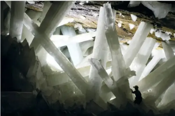 Fotografía del interior de la cueva de Naica con enormes cristales blancos de selenita; una persona diminuta aparece al pie de uno de ellos, lo que permite apreciar su escala gigantesca.
