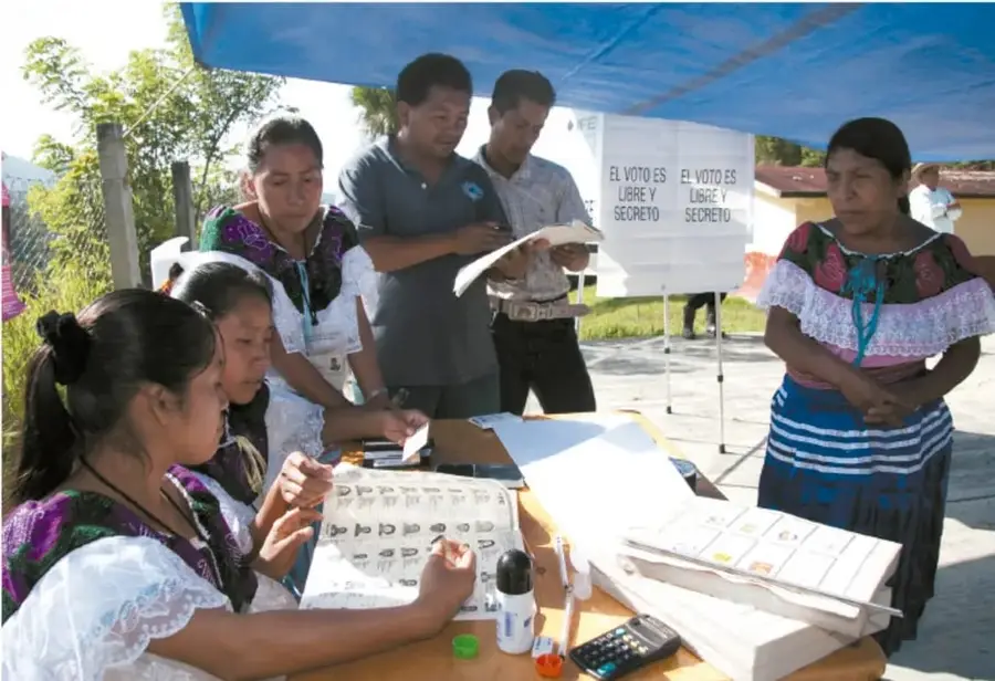 Fotografía de una casilla electoral en una comunidad rural mexicana: funcionarias de mesa con vestimenta tradicional (huipiles) atienden a votantes junto a mesas con listas nominales, calculadora y tinta; pancartas al fondo dicen 'EL VOTO ES LIBRE Y SECRETO'; bajo una lona azul, personas en fila.