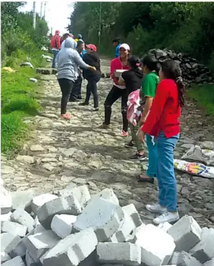 Fotografía de un grupo de personas (adultos y niñas) trabajando en comunidad en un camino empedrado rodeado de vegetación; algunas cargan o apilan piedras y materiales de construcción, ilustrando una tarea colectiva o faena propia de la ayuda mutua.