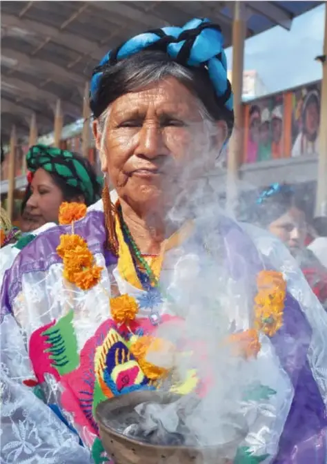 Fotografía de una mujer indígena mayor con rebozo y collar, portando un recipiente con flores y humo (sahumerio), posiblemente en una ceremonia o mercado; al fondo otras personas y textiles.