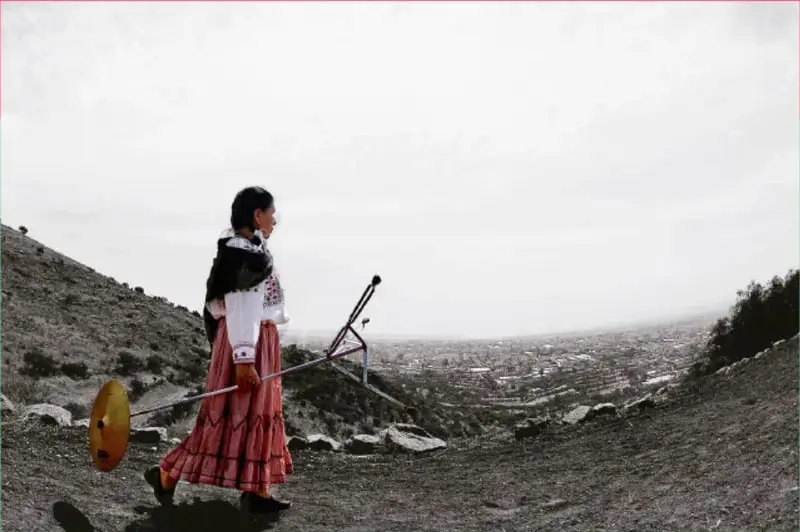 Fotografía de una niña de perfil con vestido tradicional rosado y rebozo negro, caminando por un camino de cerro con una herramienta de labranza al hombro, con una vista panorámica de la ciudad en blanco y negro al fondo. Acreditada a Raúl Barajas, Ciudad de México.