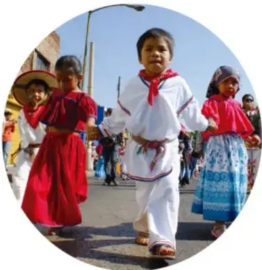Niñas y niños con trajes típicos mexicanos bailando en una calle durante una celebración cultural.