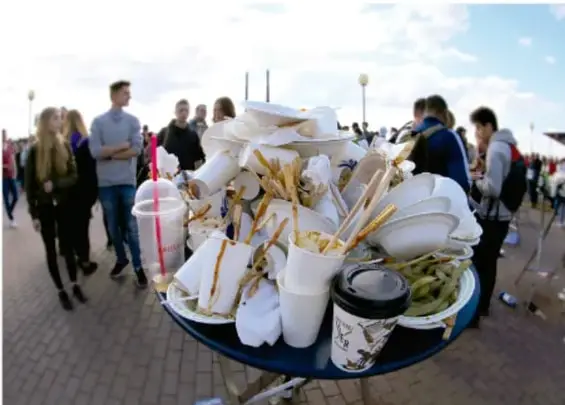 Fotografía de una mesa con desechos de comida y vasos desechables desbordados en un espacio público con personas al fondo, ilustrando una situación de riesgo sanitaria y ambiental.