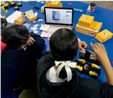 Fotografía de estudiantes trabajando en equipo con una computadora portátil y materiales amarillos sobre una mesa azul, diseñando un prototipo.