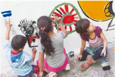 Niñas y niños pintando con brochas un mural colorido en una pared.