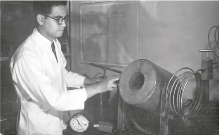 Fotografía en blanco y negro de Luis Ernesto Miramontes Cárdenas en un laboratorio, con bata blanca y lentes, manipulando equipo científico (un tambor metálico con espiral y un manómetro).