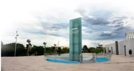 Fotografía de una plaza o espacio público con una torre de cristal y personas caminando.