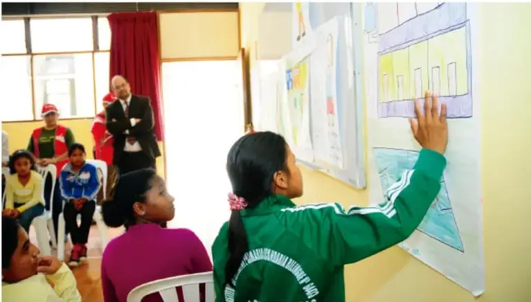 Fotografía de una estudiante de espaldas, con uniforme verde, colocando un cartel con dibujos en un periódico mural dentro del aula; sus compañeros y un adulto observan sentados.