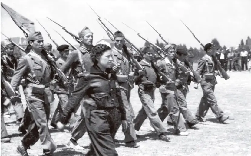 Fotografía en blanco y negro de una formación militar. Varios soldados marchan en columna con fusiles al hombro y bayonetas caladas. Al frente, una figura femenina con uniforme oscuro avanza con determinación. Al fondo se observan más tropas y un paisaje abierto.