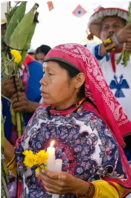 Fotografía de una ceremonia comunitaria: mujer indígena con rebozo y vestimenta tradicional sosteniendo una vela encendida y flores amarillas; detrás, un hombre con sombrero tradicional sostiene plantas de maíz y otra persona toca un instrumento musical.