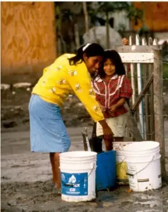 Fotografía a color de una mujer y una niña llenando cubetas de agua en una llave comunitaria.