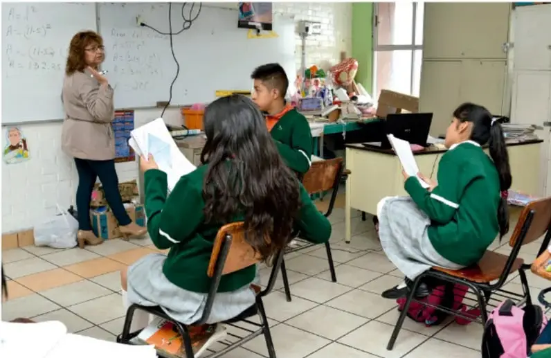 Fotografía de un salón de clases donde una docente y estudiantes en uniforme verde discuten en comunidad, revisando hojas de trabajo; se observa pizarrón con anotaciones y materiales escolares.