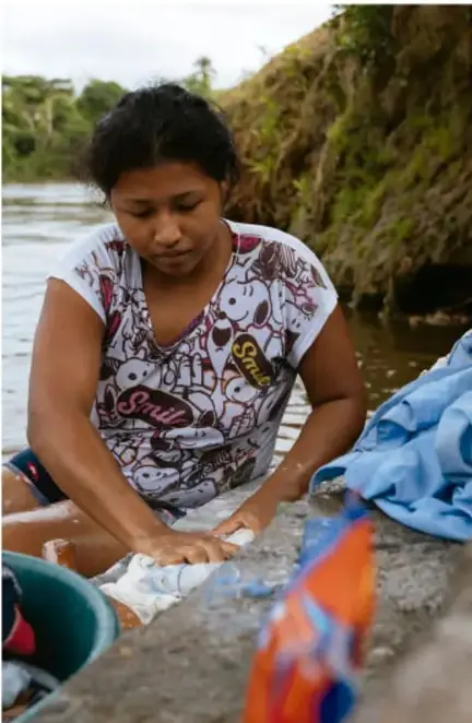 Fotografía de una joven indígena lavando ropa a orillas de un río, rodeada de vegetación; ilustra la vida cotidiana de comunidades hablantes de lenguas indígenas.