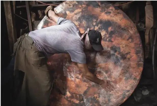 Artesano trabajando una gran pieza circular de cobre martillándola en un taller.