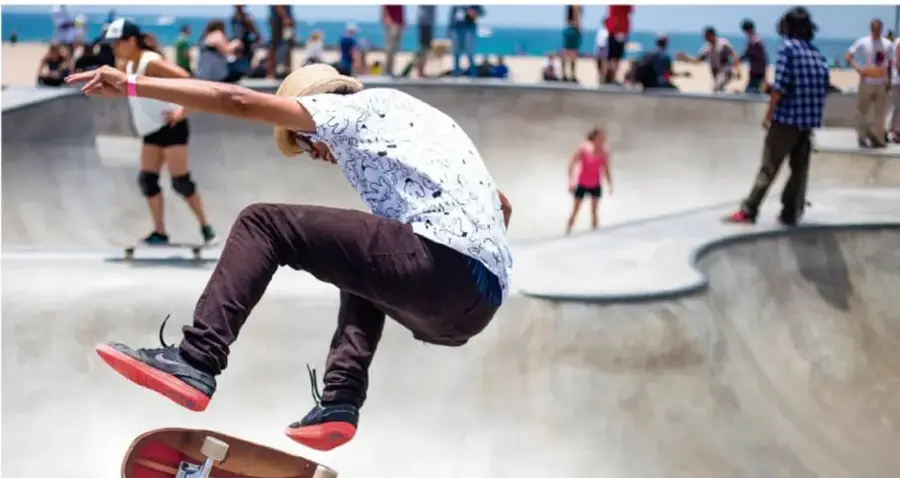 Fotografía de un joven con sombrero haciendo una maniobra de skateboard en una pista de skate al aire libre junto a la playa; al fondo hay otros jóvenes y personas observando.