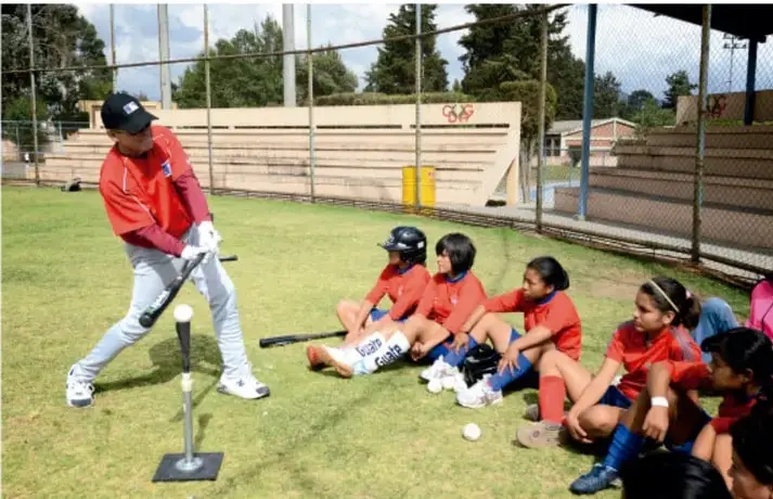Instructor con guante y bate mostrando la técnica de bateo a un grupo de alumnos con uniforme rojo sentados en el pasto de una cancha con gradas y cercas.