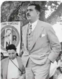 Fotografía en blanco y negro del general Lázaro Cárdenas con traje y corbata, acompañado de una persona; al fondo se ve un cartel con las siglas PNR.
