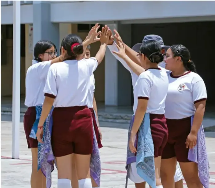 Grupo de estudiantes con uniforme escolar (camiseta blanca y pantalón corto vino) dando seña de unión con las manos alzadas en el patio de la escuela, símbolo de colaboración y trabajo en equipo.