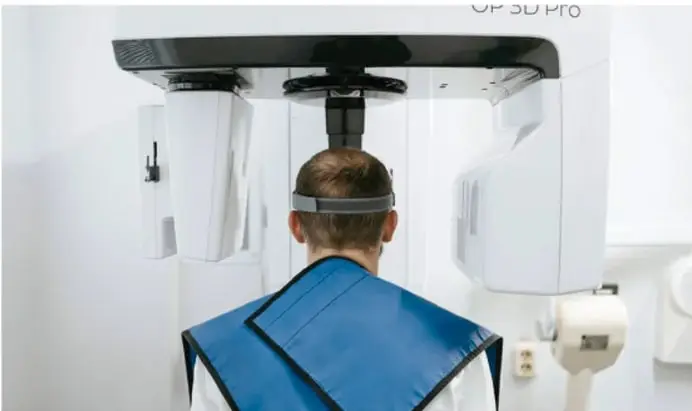 Fotografía de un paciente sentado frente a un equipo de rayos X dental (marca 'OR SD PRO'), con delantal plomado azul y soporte para la cabeza, mientras se le realiza un estudio radiográfico.