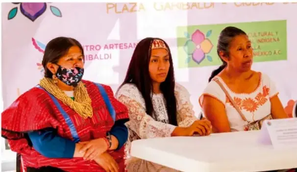 Fotografía de tres mujeres indígenas vestidas con trajes tradicionales coloridos sentadas en una mesa durante un evento cultural en la Plaza Garibaldi, Ciudad de México.