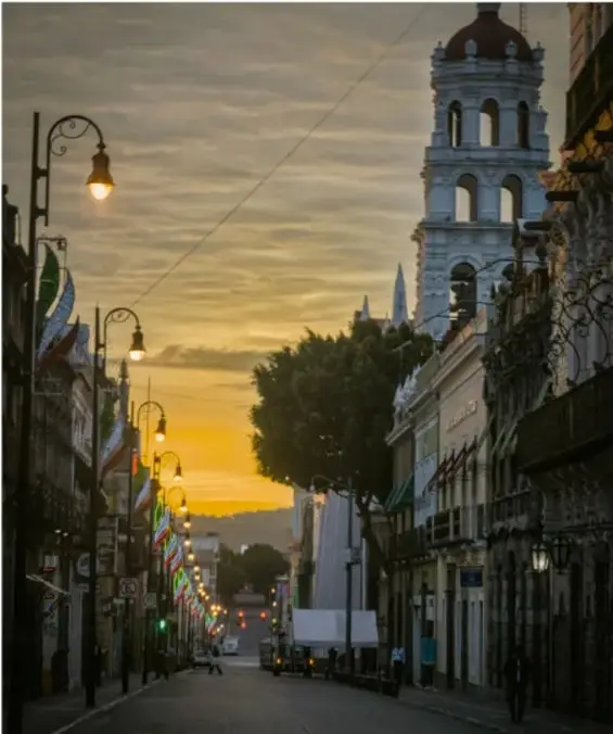 Fotografía de una calle de un pueblo mexicano al atardecer, con faroles encendidos, una torre de iglesia y banderines de colores.