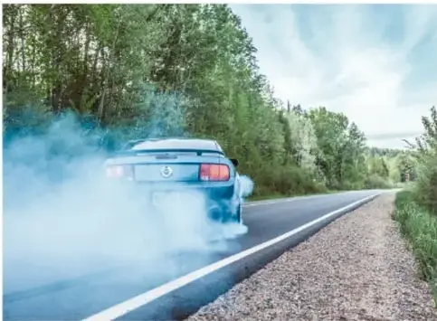Automóvil azul circulando por una carretera con abundante humo blanco saliendo del escape, rodeado de árboles.