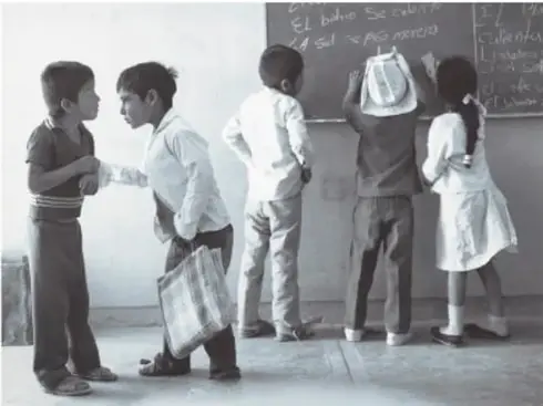 Fotografía en blanco y negro de niños dentro de un salón: dos platican entre sí y otros dos escriben en un pizarrón; La sal se puso morena, 1989, Puebla, de Mariana Yampolsky (1925-2002).