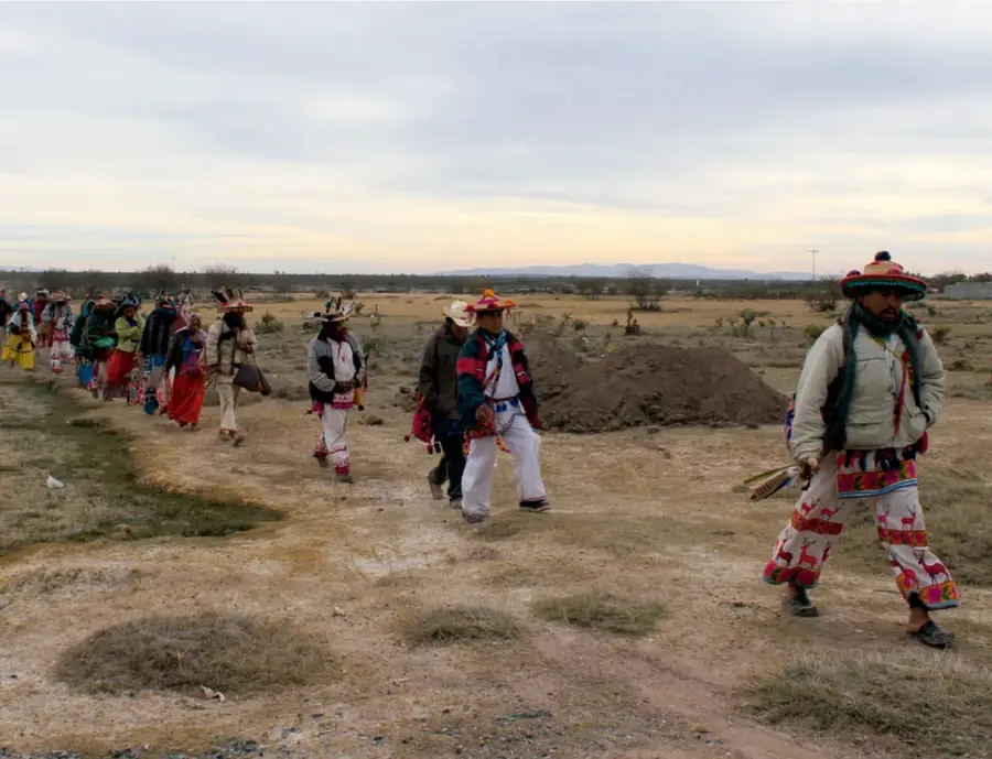 Fotografía de una procesión de personas con vestimenta tradicional indígena (sombreros bordados, calzas y ropas de colores) caminando por un camino de tierra en un paisaje árido y abierto al atardecer.