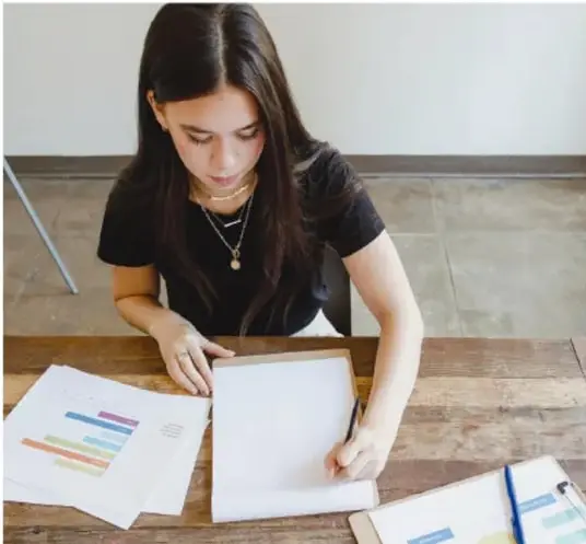 Fotografía de una estudiante sentada en una mesa de madera, revisando y organizando hojas impresas con gráficas de colores y notas adhesivas, durante la preparación de su proyecto.