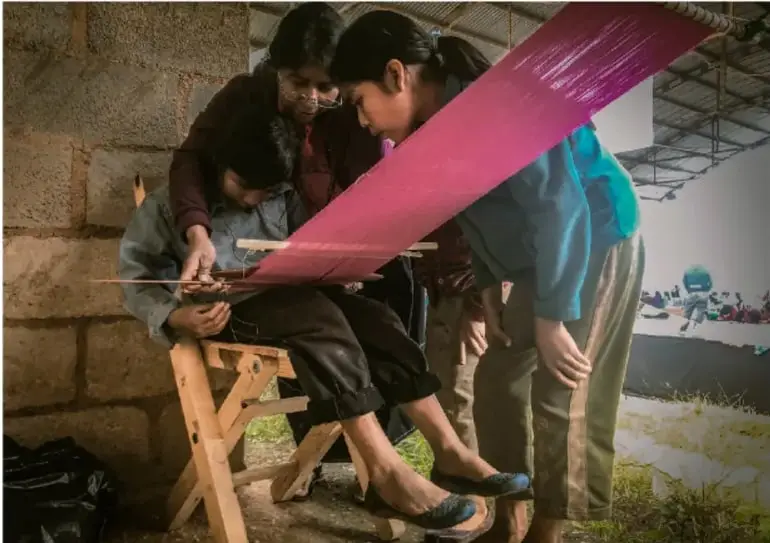 Fotografía de personas jóvenes tejiendo en un telar con hilos rosas, en un espacio comunitario, ilustrando prácticas culturales propias.
