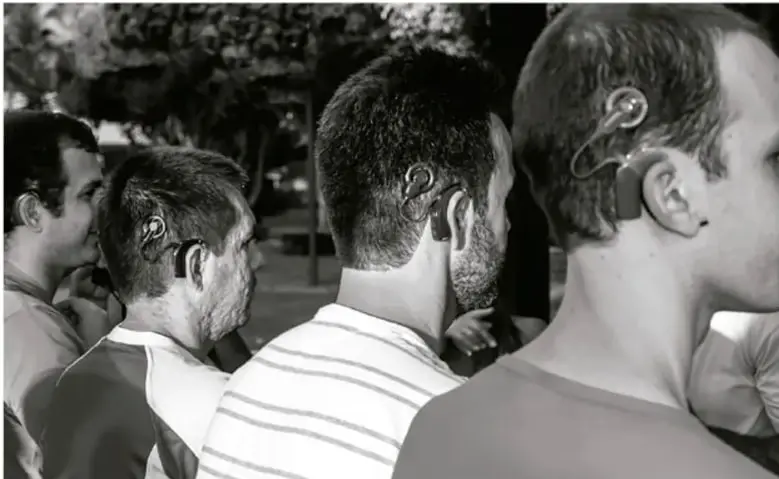Fotografía en blanco y negro que muestra de perfil a cuatro hombres; dos de ellos llevan un dispositivo auditivo (implante o audífono) detrás de la oreja, ilustrando la tecnología coclea mencionada en el texto.