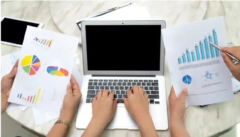 Cuatro personas alrededor de una mesa trabajan con una laptop y hojas impresas con gráficas de barras y circulares; una mano señala un gráfico con un lápiz.